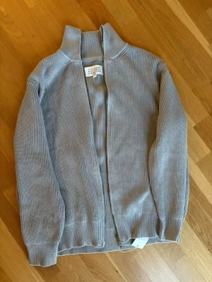 Ljusgrå stickad ziptröja Maison Margiela - Säljer en ljusgrå stickad ziptröja från Maison Margiela. Tröjan har hög krage, ribbade muddar och dragkedja framtill. Perfekt för lager på lager och enkel att matcha med olika stilar.