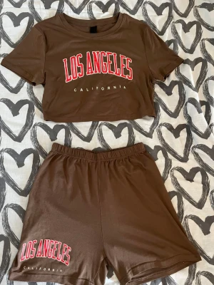 Klädset - Tvådelat fint skönt set i brunt med croppad t-shirt och shorts. T-shirten har tryck med texten 'LOS ANGELES CALIFORNIA' i rött och vitt. Shortsen har matchande tryck på ena benet och elastisk midja. Mjukt och bekvämt material, perfekt för chill dagar.