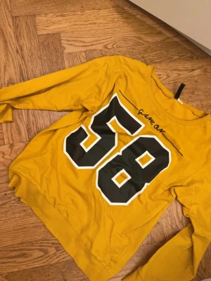 Gul långärmad topp med tryck - Cool gul långärmad topp med stort svart och vitt tryck av siffran 58 på ryggen och texten 'Game On' ovanför. Perfekt för dig som gillar sportig stil och vill sticka ut. Skön passform och mjukt material.