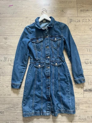 Jeansklänning från Vero Moda - Säljer en snygg jeansklänning från Vero Moda med långa ärmar, knappar framtill och fickor på bröstet. Klänningen har markerad midja och klassisk skjortkrage. Perfekt för dig som gillar denim och vill ha en cool vardagslook. Har inga skavanker.