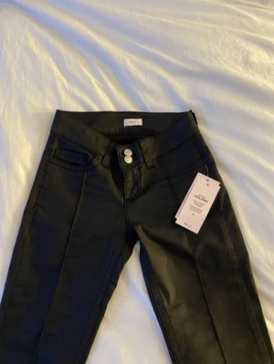 Nelly svarta coated jeans  - Snygga svarta coated jeans från Nelly med låg midja och utsvängda ben. Jeansen har dubbla silverknappar i midjan, klassiska fickor fram och bak samt snygga sömmar på bakfickorna, nästintill oanvända💕