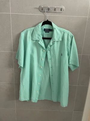 Mintgrön kortärmad skjorta Polo Ralph Lauren - Fräsch mintgrön skjorta från Polo Ralph Lauren med korta ärmar och klassisk krage. Skjortan har knappar framtill och den ikoniska loggan broderad på bröstet. Perfekt för en avslappnad och stilren look. Kanske lite skrinklig men det är för jag har typ aldrig andvänt den och den har legat i garderoben i ett tag