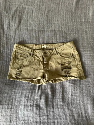 Jeansshorts med låg midja - Säljer ett par beige/grön/bruna jeans shorts med låg midja och slitna detaljer. De är perfekta för sommaren.