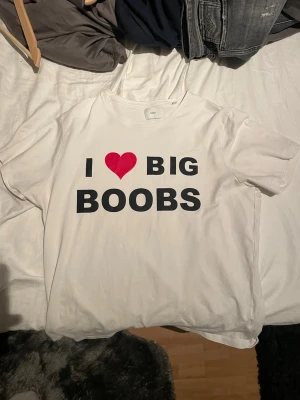Vit t-shirt med tryck och hjärta - Vit t-shirt med svart text och ett rött hjärta på bröstet. Trycket säger 'I ❤️ BIG BOOBS' 