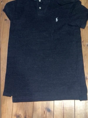 Svart pikétröja från Ralph Lauren - Klassisk svart pikétröja från Ralph Lauren med korta ärmar och vit broderad logga på bröstet. Tröjan har krage och knäppning med två knappar framtill. Perfekt till en avslappnad men ändå stilren look.