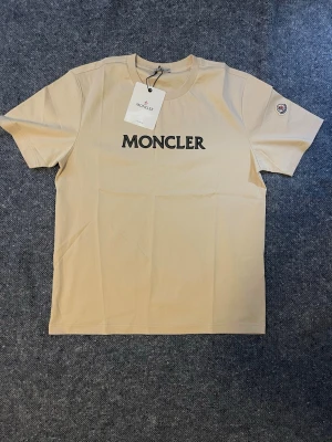 Beige Moncler t-shirt med logga - Snygg beige t-shirt från Moncler med svart logotyp tryckt på bröstet och Moncler-märke på ärmen. Klassisk passform och rund halsringning. Perfekt basplagg för en clean och stilren look. Finns även i navy blå och svarta. Storlek L