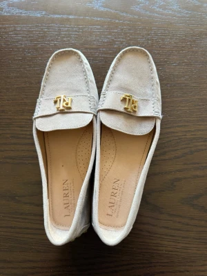 Beige loafers från Lauren Ralph Lauren - Snygga beige loafers i mocka från Lauren Ralph Lauren med guldfärgade RL-detaljer framtill. Skorna har klassisk form, mjuk innersula och gummisula med nitar för extra grepp. Perfekta för en stilren och avslappnad look.