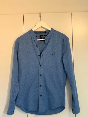Blå rutig henley-skjorta Hollister - Blå henley-skjorta från Hollister med diskret rutigt mönster och mörka knappar. Skjortan har lång ärm, rundad halsringning utan krage och en liten broderad logga på bröstet. Tillverkad i mjuk bomull för en bekväm känsla. Priset är inte hugget i sten!!