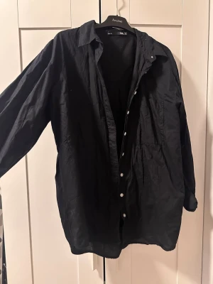 Svart skjorta - Klassisk svart skjorta med vita knappar och bröstficka. Skjortan har lång ärm och avslappnad passform, perfekt för en stilren look. Tillverkad i bomull för en skön känsla.