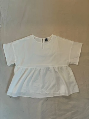 Ny Vit blus med volang från SHEIN - Helt ny vit blus från SHEIN i storlek XS. Blusen har rund halsringning, korta ärmar och en volangdetalj nedtill som ger en luftig och avslappnad look. Perfekt att styla med jeans eller kjol för en trendig outfit.