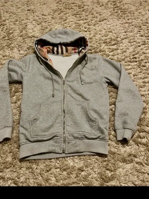 Grå zip-hoodie från Burberry - Snygg grå hoodie från Burberry med dragkedja och klassiskt rutigt foder i huvan. Mjuk bomullskänsla, två stora fickor fram och justerbar dragsko i huvan. Perfekt för en chill och stilren look. Inga fläckar helt ny. Var för liten det är därför jag säljer den! Ifall de är frågor ni undrar så är de bara skriva!!
