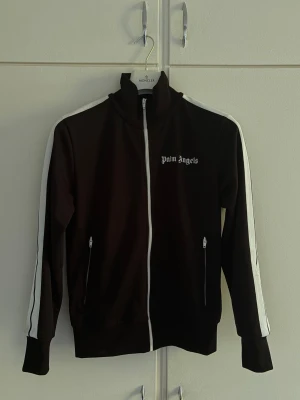 Svart track jacket från Palm Angels (Äkta) - Svart track jacket från Palm Angels med vit dragkedja och vita ränder längs ärmarna. Hög krage och två fickor med dragkedja framtill. Ikonisk logga på bröstet. Äkta och kvitto finns. 