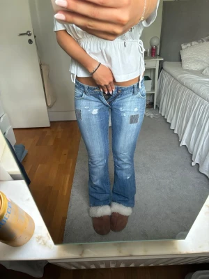 Ljusblå bootcut jeans med slitningar - Säljer ett par ljusblå bootcut jeans med låg midja och slitna detaljer framtill. Jeansen har patchade partier och en avslappnad passform. Perfekta för en trendig och avslappnad stil.