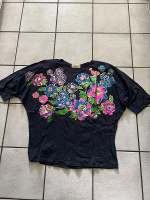 Svart t-shirt med färgglada blommor storlek XL bomull  - Unik svart t-shirt med stort färgglatt blommönster i blått, rosa, lila och grönt över bröstet och axlarna. T-shirten har korta ärmar och en loose passform, perfekt för dig som gillar statement-plagg och vill sticka ut.