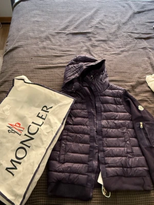 Marine blue Moncler Cardigan med NFC - Snygg lila pufferjacka från Moncler med huva och stickade ärmar. Jackan har dragkedja framtill, två fickor och en extra ficka med dragkedja på ärmen. Moncler-logga på armen och ribbade muddar vid ärmslut och nederkant. Perfekt för kalla vinterdagar.
