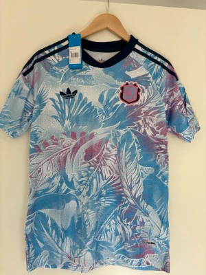 Adidas Costa Rica  sporttröja M - Säljer en snygg sporttröja från Adidas med blått, vitt och lila tropiskt bladmönster. Tröjan har mörkblå ribbad krage, svarta ränder på axlarna och Adidas-logga på bröstet. Tillverkad i lätt och ventilerande material, perfekt för träning eller streetwear.