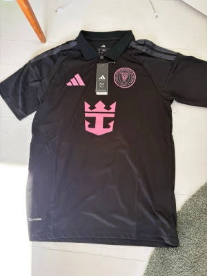 Svart Inter Miami matchtröja Adidas - Snygg svart matchtröja från Inter Miami CF i slim fit-modell från Adidas. Tröjan har rosa detaljer, klubbmärke och sponsorlogga på bröstet samt klassisk pikékrage. Tillverkad i lätt och ventilerande material med Climacool-teknologi.