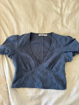 Blå broderad blus från NAKD - Snygg blå blus från NAKD med broderade blommor och puffärmar. Toppen har v-ringning och är croppad, vilket ger en trendig look. Perfekt till jeans eller kjol för en cool outfit.