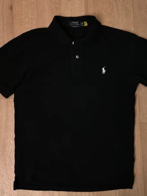 Svart pikétröja från Polo Ralph Lauren - Klassisk svart pikétröja från Polo Ralph Lauren med vit broderad logga på bröstet. Tröjan har korta ärmar, krage och knappar framtill. Tillverkad i mjuk bomull för en bekväm känsla och stilren look.