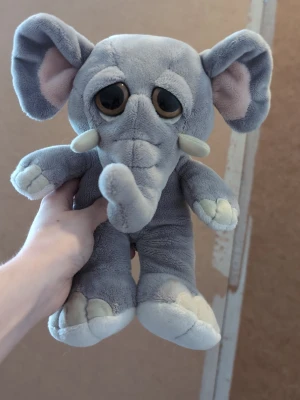 Gosedjur elefant med stora öron - Mjuk och gosig elefant i plysch med stora bruna ögon, grå kropp och ljusa detaljer på fötter, betar och insidan av öronen. Elefanten har en lång snabel, stora öron och liten svans. Perfekt som kramkompis eller dekoration.