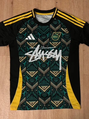 Jamaica x Stüssy x Adidas matchtröja - Unik Jamaica matchtröja i samarbete mellan Adidas och Stüssy. Svart bas med grönt och gult geometriskt mönster, gula detaljer och Stüssy-logga framtill. Märkt med 'Reggae Boyz' i nacken och Jamaica-emblem på bröstet. Tillverkad i lätt funktionsmaterial.