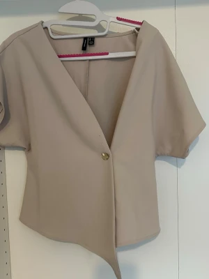 Beige kortärmad blus  - Stilren beige blus med korta ärmar och djup V-ringning. Blusen har en asymmetrisk nederkant och stängs med en dekorativ guldfärgad knapp framtill. 