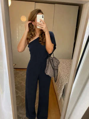 Mörkblå one shoulder jumpsuit Lauren Ralph Lauren - Snygg mörkblå jumpsuit från Lauren Ralph Lauren i elegant one shoulder-modell. Vida ben och cape-liknande detalj över ena axeln. Tillverkad i mjukt georgette-tyg som ger ett lyxigt fall. Perfekt för dig som vill sticka ut med stilren look.