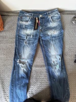 Distressed jeans från Dsquared2 - Säljer ett par blå distressed jeans från Dsquared2 med slitningar, hål och färgstänk för en edgy look. Klassisk femficksmodell med dragkedja och patch bak i midjan med 'CaTEN'. Perfekta för dig som gillar streetwear och statement-plagg.