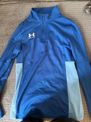 Blå träningströja från Under Armour - Snygg blå långärmad träningströja från Under Armour med halv dragkedja och ljusblå paneler på sidorna. Perfekt för löpning eller gym. Tillverkad i ett mjukt och stretchigt funktionsmaterial som andas bra. Logga på bröstet.