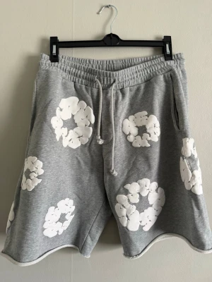 Gråa Denim Tears shorts - Grå mjuka shorts från Denim Tears med vita blommiga tryck över hela plagget. Shortsen har resårmidja med snörning och sidofickor. Perfekta för chill dagar och har en relaxed fit som ger skön känsla. Helt nya aldrig använda i storlek L perfekt nu för sommarn