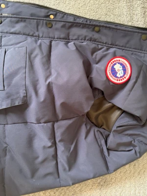 Mörkgrå dunjacka från Canada Goose - Säljer en mörkgrå dunjacka från Canada Goose med klassiskt rundt märke på ärmen. Jackan har stora fickor framtill och robusta tryckknappar längs kanten. Materialet är slitstarkt och fodret är mjukt, perfekt för kalla vinterdagar.