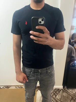Svart t-shirt från Polo Ralph Lauren - Svart t-shirt från Polo Ralph Lauren med klassisk röd logga broderad på bröstet. T-shirten har rund halsringning och korta ärmar. Tillverkad i mjuk bomull för en skön och stilren look.