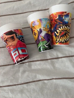 Färgglada plastmuggar med motiv - Tre plastmuggar med olika färgstarka motiv inspirerade av tecknade serier och sport. Muggarna har vita lock och tryck i rött, blått, gult, lila och orange. Perfekta för dryck eller som dekoration på skrivbordet.