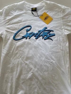 Vit t-shirt från Corteiz med blå logga - Snygg vit t-shirt från Corteiz med stor blå och svart logga i graffiti-stil på bröstet. Klassisk passform och rund halsringning. Perfekt för dig som vill ha en clean men ändå statement look.