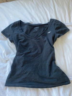 Svart v-ringad t-shirt Hollister - Snygg svart t-shirt från Hollister med v-ringning och liten logga på bröstet. Klassisk passform och korta ärmar, perfekt till jeans eller kjol. Tillverkad i mjuk bomull som känns skön mot huden.