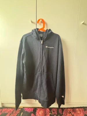 Mörkblå zip hoodie från Champion - Snygg mörkblå hoodie från Champion med dragkedja framtill och broderad logga på bröstet. Klassisk modell med huva och känguruficka. Perfekt för en avslappnad streetwear-look. Mjuk bomullsblandning som känns skön mot huden.