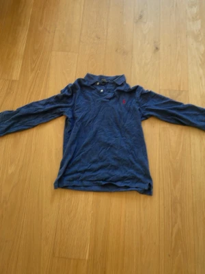 Blå långärmad polotopp Ralph Lauren - Klassisk blå långärmad polotopp från Polo Ralph Lauren i slim fit-modell. Toppen har krage, knappar framtill och den ikoniska röda loggan broderad på bröstet. Perfekt för en stilren och avslappnad look.