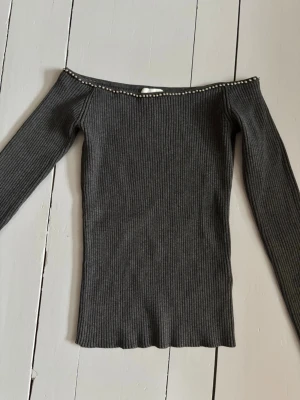 Mörkgrå off shoulder tröja med nitar - Snygg mörkgrå ribbstickad tröja med off shoulder-modell och dekorativa små nitar/ kulor längs halsringningen. Långa ärmar och figurnära passform, perfekt för en cool outfit!