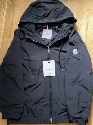 Svart vindjacka från Moncler - Snygg svart vindjacka från Moncler med huva och dragkedja framtill. Jackan har Moncler-logga på ärmen, justerbar dragsko i huvan och elastiska muddar vid ärmslut och nederkant. Tillverkad i lätt och slitstarkt material, perfekt för blåsiga dagar.