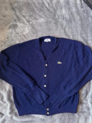 Mörkblå kofta från Lacoste - Klassisk mörkblå kofta från Lacoste med V-ringning och knäppning framtill. Koftan har långa ärmar, ribbade muddar och det ikoniska krokodilmärket broderat på bröstet. Tillverkad i mjukt material som känns skönt mot huden.