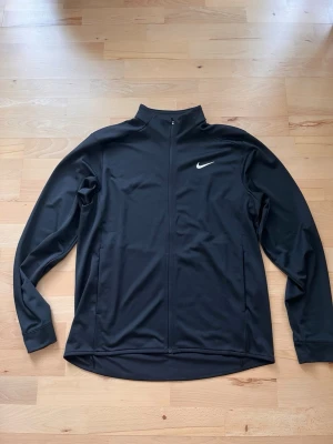 Svart träningsjacka från Nike - Säljer en svart träningsjacka från Nike med hel dragkedja och hög krage. Jackan har diskret vit logga på bröstet och praktiska fickor med tryckknapp. Materialet är lätt och stretchigt, perfekt för sport och aktivitet.