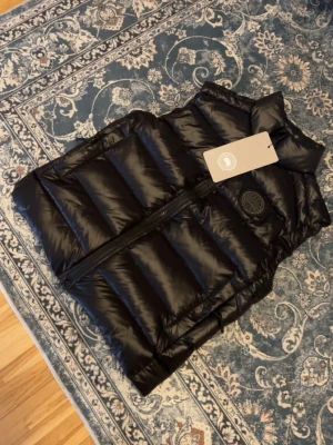 Svart dunväst från Canada Goose - Snygg svart dunväst från Canada Goose med hög krage och dragkedja framtill. Västen har ett klassiskt quiltat mönster och en rund logotyp på bröstet. Tillverkad i glansigt material och fylld med dun för extra värme. Perfekt för lager på lager under kyliga dagar.