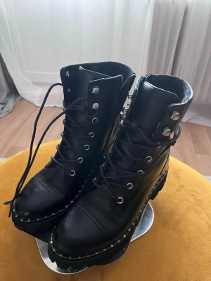 Svarta boots med nitar och snörning - Tuffa svarta stövletter i skinnimitation med grov sula och silverfärgade nitar längs sulkanten. Högt skaft med snörning och metallöljetter ger en edgy look. Perfekta för dig som gillar en rockig stil och vill sticka ut.