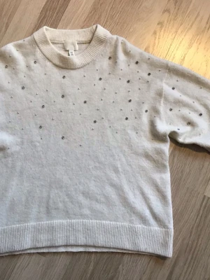 Vit stickad tröja med pärlor från H&M, stl XS - Vit stickad tröja från H&M med rund halsringning och dekorativa små pärlor och ”ädelstenar” framtill. Tröjan har ribbade muddar vid ärmslut och nederkant samt en avslappnad passform. Väldigt lite använd, så jättebra skick. Just nu fri frakt, så du betalar bara för tröjan. Jag har fler plagg ute, och hittar du något mer från min garderobsutrensning, så skickar jag i samma paket.