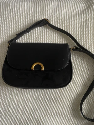 Svart axelväska med gulddetalj - Snygg svart axelväska i mocka och läder med justerbart axelband. Väskan har en elegant guldfärgad metallspänne framtill och rundad form. Perfekt för dig som gillar stilrena accessoarer med en touch av lyx.