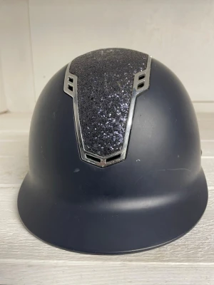 Mörkblå ridhjälm från Jacson  - Snygg mörkblå ridhjälm med en glittrig panel framtill och silverfärgade detaljer. Hjälmen har en klassisk rundad form och skärm framtill. Materialet är hård plast med dekorativ yta på ovansidan. Den är i storlek S-M, 55-57cm. Det är från Jacson och ordinarie priset är 1499kr.