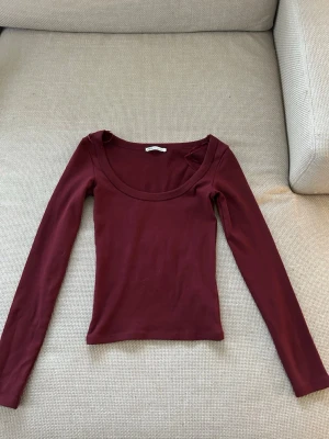 Vinröd långärmad topp från Zara - Snygg vinröd långärmad topp från Zara med rundad, bred urringning. Tillverkad i mjukt material som sitter tight mot kroppen. Perfekt att styla med jeans eller kjol för en trendig look.