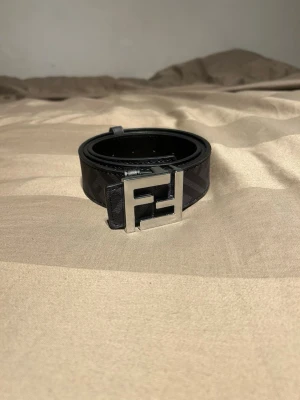 Svart Fendi bälte - Snyggt svart bälte från Fendi med det klassiska FF-mönstret längs hela bandet och ett stort, silverfärgat FF-spänne framtill. Perfekt accessoar för att lyfta vilken outfit som helst med en lyxig touch och ikonisk design.