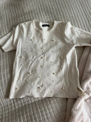 Vit topp med pärlor från Zara - Supersöt vit topp från Zara med korta ärmar och rund hals. Toppen är dekorerad med pärlor över hela framsidan och baksidan för en extra stylish look. Perfekt att styla med jeans eller kjol för en trendig outfit. Står storlek M men passar mig som bär Xs/S. 