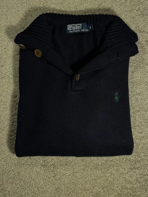 Svart stickad tröja från Polo Ralph Lauren - Svart stickad tröja från Polo Ralph Lauren med klassisk krage och knappar på axeln. Tröjan har ett broderat grönt Polo-logotyp på bröstet och är tillverkad i ett tjockt, mysigt material. Perfekt för dig som gillar stilrena och tidlösa plagg.